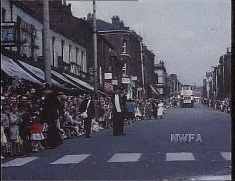 A colour photo of a busy street scene, with lots of people lining the road. There are shop awnings above them on the left size of the road, and a zebra crossing in the foreground. The letters NWFA are printed on the photograph, on an empty space in the road.